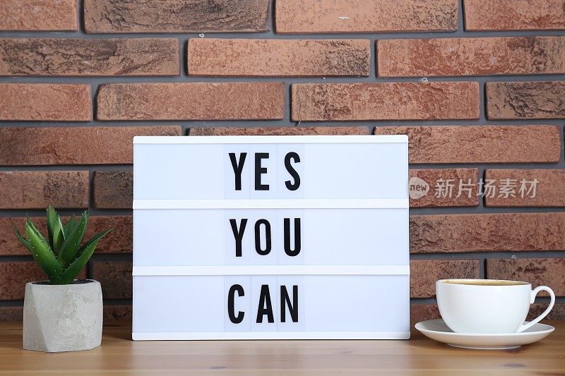 灯箱上写着“Yes You Can”，砖墙旁的桌子上放着一杯咖啡和盆栽室内植物。励志名言